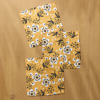 Three square dishclothes with a yellow floral pattern, laid out on a light wood surface.