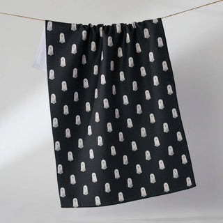 A black Geometry Tea Towel with a repeating white ghost pattern, hanging from a thin wire against a plain light gray wall.