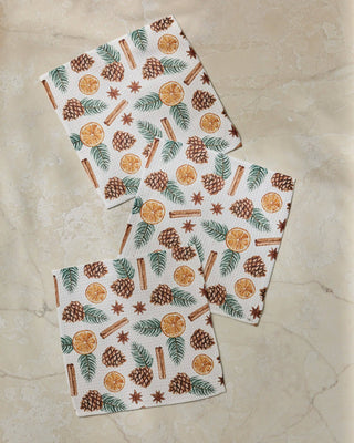 Three square white dishclothes with a festive holiday print of pinecones, cinnamon sticks, star anise, green pine needles, and orange slices, arranged on a beige marble surface.
