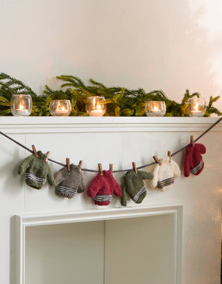 A festive holiday mantel decorated with green pine garland, lit candles in glass holders, and a string of small knit mittens hanging from wooden clips, with a glowing Christmas tree on the right.