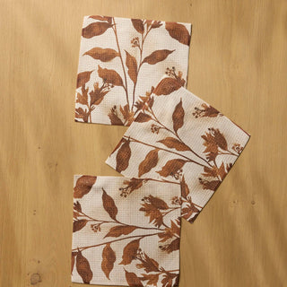 Three square dishclothes with a brown leaf and branch pattern on a cream background, arranged on a light wood surface.