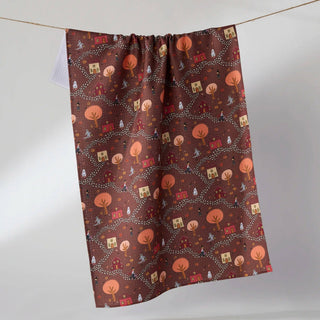 A brown tea towel with an autumn-themed pattern of orange pumpkins, small trees, and houses, hanging from a string against a light gray background.
