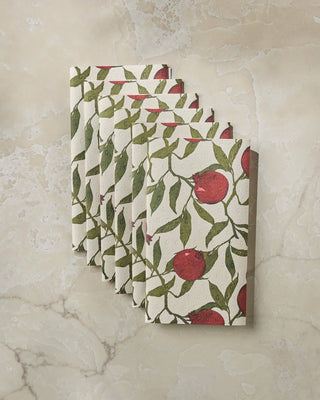 A stack of rectangular paper napkins with a botanical pattern of red pomegranates and green leaves on a cream background, displayed against a beige marble-textured wall.