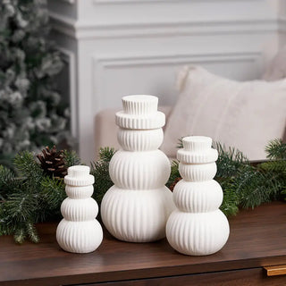 Three white ribbed snowman-shaped decorative vases arranged on a wooden surface with evergreen branches and a pinecone, in front of a softly blurred indoor background.