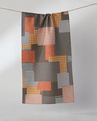 A mini towel with a patchwork print of geometric blocks in orange, brown, black, white, and gray patterns, hanging from a thin horizontal rope against a light gray wall.
