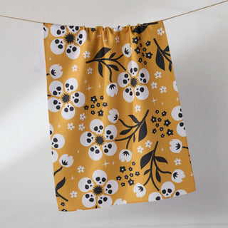 A mustard yellow kitchen towel with a white and black floral and skull pattern, hanging from a string against a plain white background.