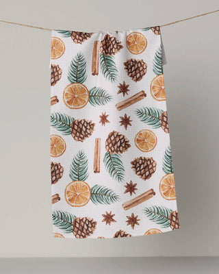 A white kitchen towel with a festive holiday print of pinecones, cinnamon sticks, star anise, green pine needles, and orange slices, hanging from a thin string against a neutral gray background.