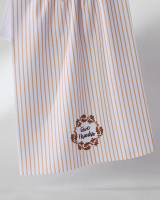 A white kitchen tea towel with thin vertical pink stripes and a brown floral wreath design featuring the words 'give thanks' in script, hanging from a string against a light gray background.