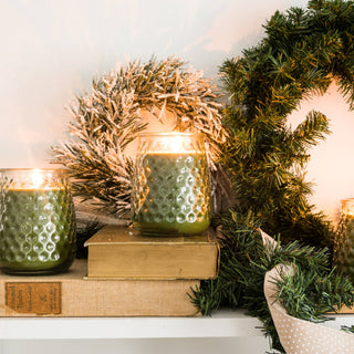 Three textured green glass jar candles with lit wicks are arranged on a white shelf alongside a green wreath, books, dried botanicals, and a cream ribbon against a white wall.