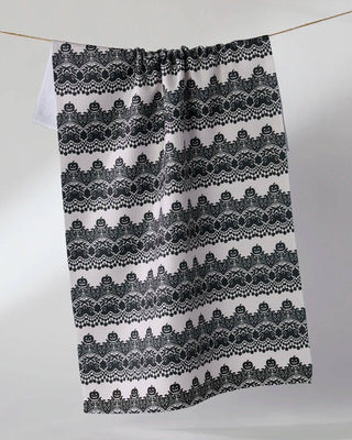 A white tea towel with a black lace pattern, hanging from a string against a light gray background.