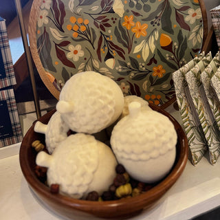 A styled tabletop display featuring a round floral-patterned serving platter, three white textured ceramic acorn ornaments in a wooden bowl, and several small patterned sachets, with a glass display case visible in the background.