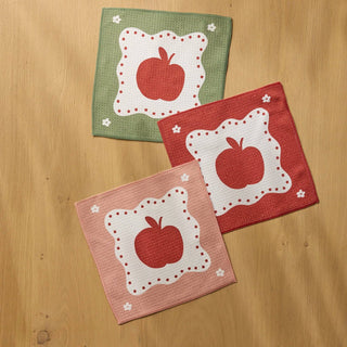 Three square dishclothes with apple designs, arranged on a light wood background. One napkin is sage green, one is red, and one is light pink, each featuring a central red apple on a white scalloped patch with small white flower motifs.