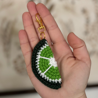 Hand holding a green and black crochet keychain with a gold clasp against a blurred background.