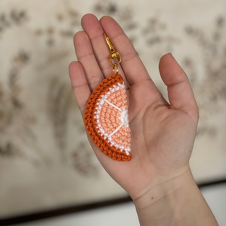 Hand holding a crocheted orange slice keychain against a neutral background