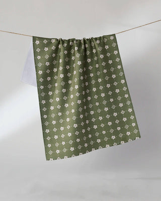 An olive green tea towel with a repeating white floral and geometric pattern, hanging from a thin wire against a neutral gray background.