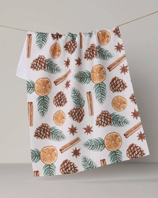 A white kitchen towel with a festive holiday print of pinecones, cinnamon sticks, star anise, evergreen branches, and orange slices, hanging from a string against a neutral gray background.