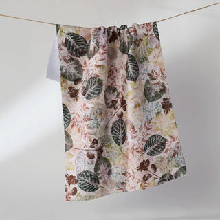 A tea towel with a botanical leaf pattern in muted green, brown, and dusty pink on a cream background, hanging from a thin wire against a plain light gray studio backdrop.