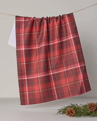 A red and black plaid kitchen towel hanging from a string against a neutral gray background, with pine branches and pinecones at the bottom right.