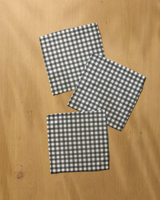 Three square black and white gingham check dishclothes arranged diagonally on a light brown wooden tabletop.