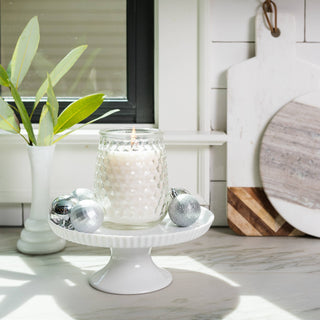 A lit white candle in a textured glass jar sits on a white pedestal stand on a kitchen counter, surrounded by silver holiday ornaments. A white vase with green leaves and rustic wooden cutting boards lean against a white subway tile backsplash in the background.