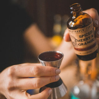 A person holding a bottle of Sweethaven Tonics with a variety of flavors, pouring the contents into a jigger.
